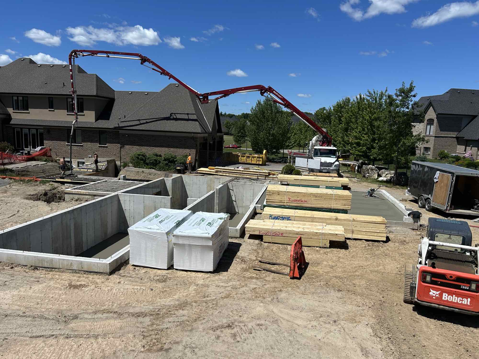 Country Estates Construction concrete work: concrete pump truck reaching across a new home foundation, with lumber and a Bobcat on site